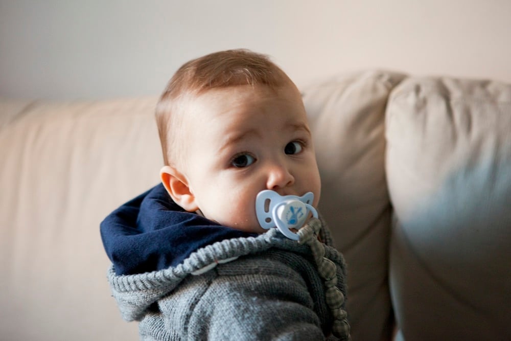 Bebé con chupete azul y jersey de punto gris sentado en un sofá de tonos claros