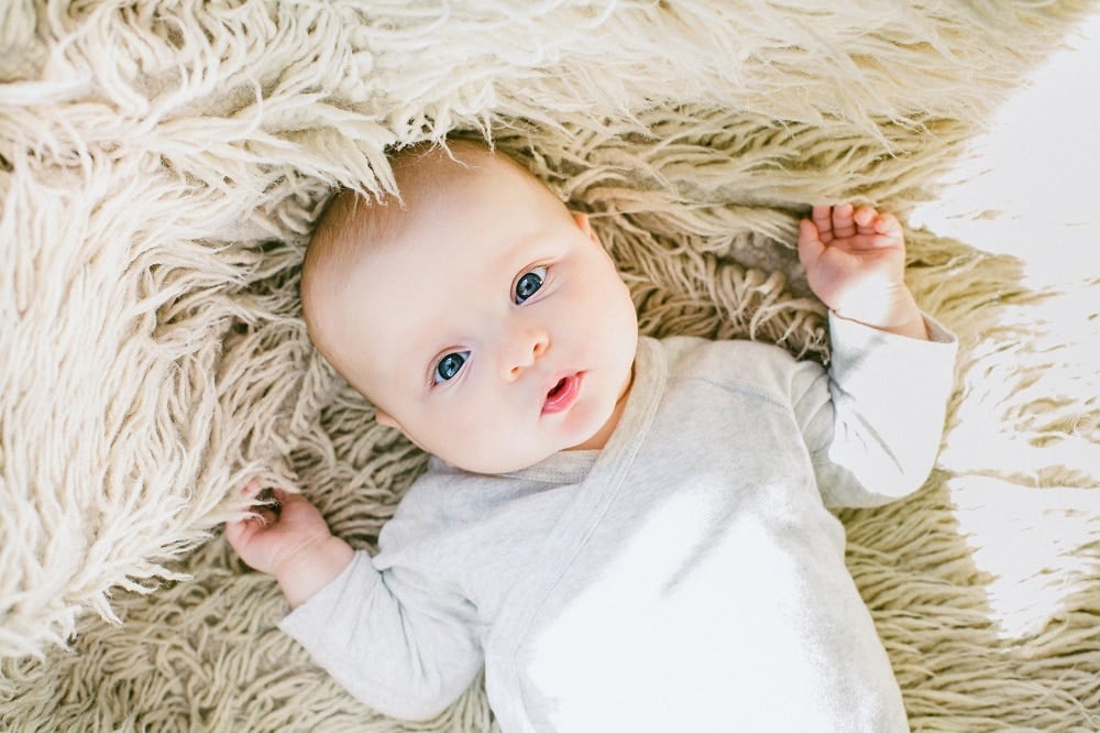 Bebé de ojos azules recostado sobre una alfombra de pelo largo blanca vistiendo ropa gris clara.