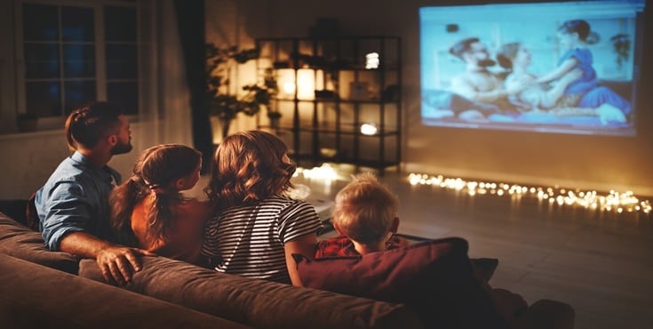 Familia en el sofá viendo una proyección de video casero en una sala acogedora iluminada con luces cálidas de suelo