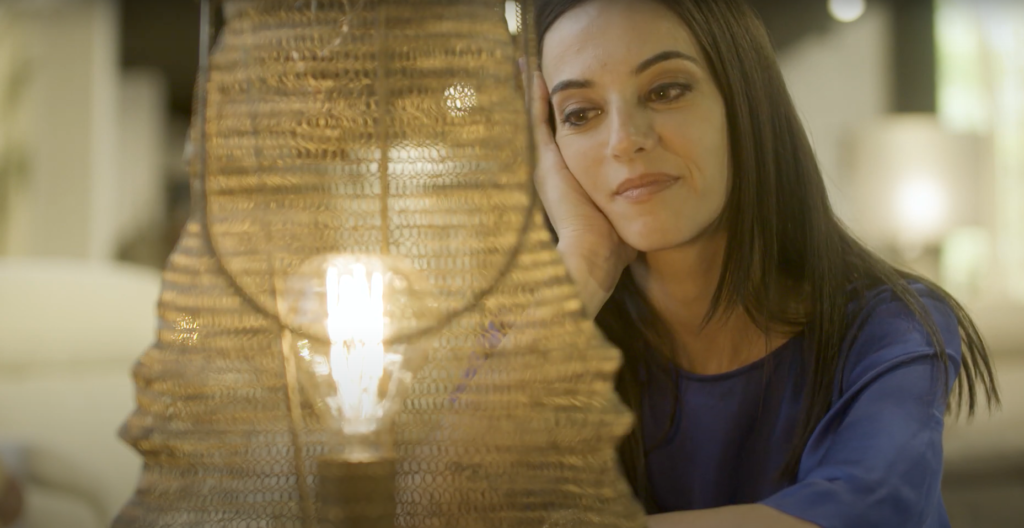 Mujer contemplando una lámpara de diseño con malla metálica dorada y bombilla de filamento encendida en un ambiente cálido