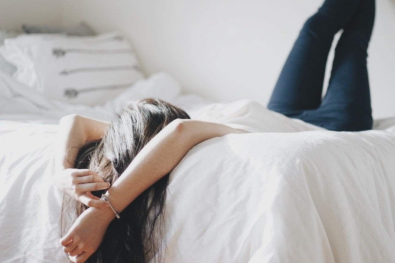 Mujer de cabello largo oscuro descansando en una cama blanca con las piernas elevadas y brazos tras la cabeza