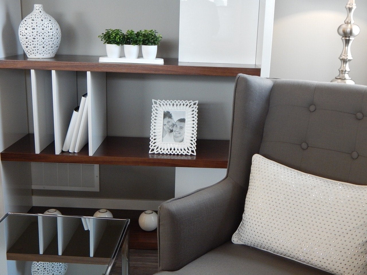 Sillón gris moderno con cojín blanco brillante junto a estantería de madera oscura y decoración minimalista en blanco