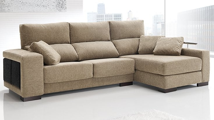 Sofá chaise longue beige con tapicería de tela y cojines decorativos en un salón moderno con grandes ventanales.